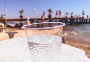 um copo de plástico está sobre a mesa, uma bebida carbonatada transparente é derramada dentro. embalagem não ecológica, copo plástico polui o meio ambiente. férias na praia foto