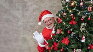 papai noel está comemorando o natal com felicidade e emoção enquanto se esconde atrás da árvore de natal totalmente decorada e acena para saudação e felicidade da temporada foto