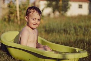 menino bonitinho tomando banho na banheira ao ar livre no jardim. criança feliz está espirrando, brincando com água e se divertindo. temporada de verão e recreação. ficar fresco no calor do verão. diversão aquática no quintal. foto