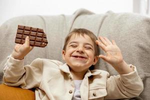 menino adorável sentado no sofá em casa e comendo barra de chocolate. criança e doces, confeitos de açúcar. criança desfrutar de uma deliciosa sobremesa. criança pré-escolar com roupas casuais. emoção positiva. foto