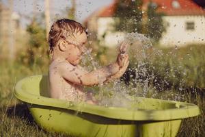 menino bonitinho tomando banho na banheira ao ar livre no jardim. criança feliz está espirrando, brincando com água e se divertindo. temporada de verão e recreação. ficar fresco no calor do verão. diversão aquática no quintal. foto