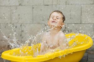 menino bonitinho tomando banho na banheira amarela ao ar livre. criança feliz está espirrando, brincando com água e se divertindo. temporada de verão e recreação. ficar fresco no calor do verão. diversão aquática no quintal. foto