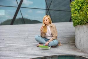 jovem linda loira comendo sushi ao ar livre, no terraço de madeira, por um edifício moderno da cidade. comida saborosa para ir. menina tem pausa para o almoço, passando o tempo fora e comendo comida asiática. vida urbana foto