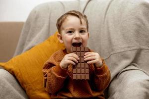 menino adorável sentado no sofá em casa e comendo barra de chocolate. criança e doces, confeitos de açúcar. criança desfrutar de uma deliciosa sobremesa. criança pré-escolar com roupas casuais. emoção positiva. foto