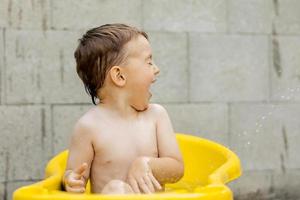 menino bonitinho tomando banho na banheira amarela ao ar livre. criança feliz está espirrando, brincando com água e se divertindo. temporada de verão e recreação. ficar fresco no calor do verão. diversão aquática no quintal. foto