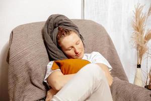 mulher jovem e loira com roupas casuais, toalha na cabeça, sentada no sofá e descansando. relaxar depois de tomar banho. autocuidado, tempo para você. relaxando em casa. mulheres, conceito de beleza e higiene. foto