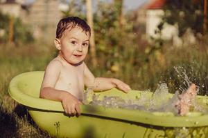 menino bonitinho tomando banho na banheira ao ar livre no jardim. criança feliz está espirrando, brincando com água e se divertindo. temporada de verão e recreação. ficar fresco no calor do verão. diversão aquática no quintal. foto