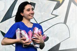 retrato de uma jovem emocional com cabelo preto e piercings. foto de uma menina com latas de tinta aerossol nas mãos em um fundo de parede de graffiti. o conceito de arte de rua e uso de tintas aerossóis