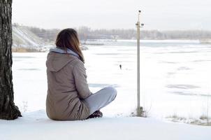 uma jovem caucasiana com um casaco marrom olhando para longe na linha do horizonte entre o céu e o lago congelado no inverno foto