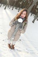 uma jovem e alegre garota caucasiana com um casaco marrom segura uma bola de neve em uma floresta coberta de neve no inverno. foto olho de peixe