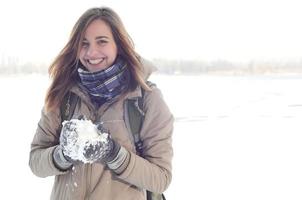 uma jovem e alegre garota caucasiana com um casaco marrom segura uma bola de neve no fundo de uma linha do horizonte entre o céu e um lago congelado no inverno foto