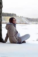 uma jovem caucasiana com um casaco marrom está sentada perto de um penhasco no fundo de uma linha do horizonte entre o céu e um lago congelado no inverno foto