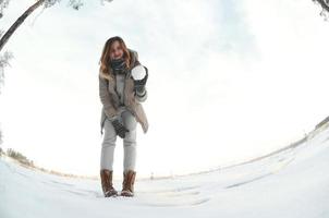 uma jovem e alegre garota caucasiana com um casaco marrom segura uma bola de neve na frente de uma linha do horizonte entre o céu e um lago congelado no inverno. foto olho de peixe