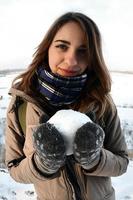 uma jovem e alegre garota caucasiana com um casaco marrom segura uma bola de neve na frente de uma linha do horizonte entre o céu e um lago congelado no inverno. foto olho de peixe