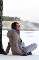 uma jovem caucasiana com um casaco marrom está sentada perto de um penhasco no fundo de uma linha do horizonte entre o céu e um lago congelado no inverno foto