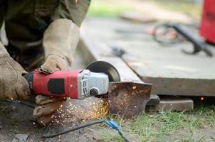 moagem de roda elétrica na estrutura de aço ao ar livre. antigo trabalhador manual em luvas de proteção cortando porta de metal com moedor foto