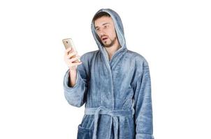 retrato de jovem barbudo caucasiano em roupão azul mostra emoção surpresa e assiste à tela do smartphone isolada no fundo branco foto