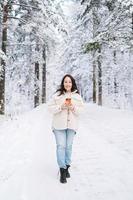 mulher sorridente com cabelo escuro em roupas de inverno com papel xícara de café nas mãos na floresta de inverno nevado foto