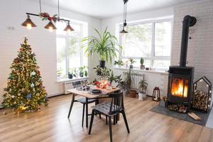interior festivo da casa é decorado para o natal e ano novo em estilo loft com fogão preto, lareira, árvore de natal. sala de estúdio quente com mesa posta, queima de madeira, aconchegante e aquecimento de casa foto