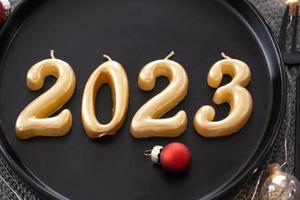 figuras douradas 2023 de velas em um prato preto em uma mesa festiva com uma porção de ano novo. interior em estilo loft, festa, banquete. guardanapo de vime, garfo, faca foto