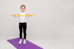 menina fica em uma esteira de ginástica e segura um bastão de ginástica com os braços estendidos na frente dela, fitness infantil, esportes foto