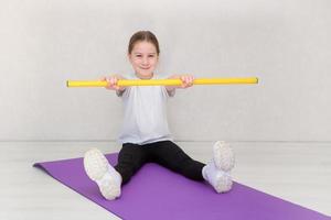 menina bonitinha senta-se em uma esteira e realiza exercícios com um bastão de ginástica sorrindo foto