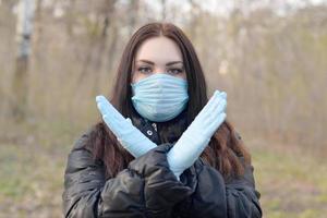 retrato de jovem morena em máscara protetora azul e luvas de borracha mostra gesto de parada ao ar livre em madeira de primavera foto