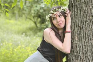 uma foto da floresta de uma bela jovem morena de aparência européia com olhos castanhos escuros e lábios grandes. na cabeça da menina está uma coroa de flores, na testa enfeites brilhantes