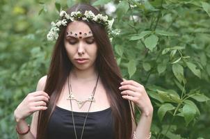 uma foto da floresta de uma bela jovem morena de aparência européia com olhos castanhos escuros e lábios grandes. na cabeça da menina está uma coroa de flores, na testa enfeites brilhantes