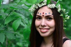 uma foto da floresta de uma bela jovem morena de aparência européia com olhos castanhos escuros e lábios grandes. na cabeça da menina está uma coroa de flores, na testa enfeites brilhantes