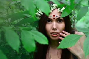 retrato de uma jovem emocional com uma coroa de flores na cabeça e enfeites brilhantes na testa. linda morena posando em uma bela floresta florescente durante o dia em um belo dia foto