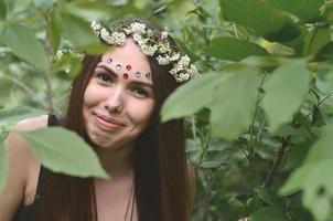 retrato de uma jovem emocional com uma coroa de flores na cabeça e enfeites brilhantes na testa. linda morena posando em uma bela floresta florescente durante o dia em um belo dia foto