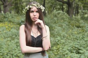 retrato de uma jovem emocional com uma coroa de flores na cabeça e enfeites brilhantes na testa. linda morena posando em uma bela floresta florescente durante o dia em um belo dia foto