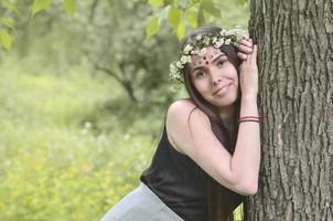 retrato de uma jovem emocional com uma coroa de flores na cabeça e enfeites brilhantes na testa. linda morena posando em uma bela floresta florescente durante o dia em um belo dia foto