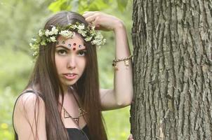 uma foto da floresta de uma bela jovem morena de aparência européia com olhos castanhos escuros e lábios grandes. na cabeça da menina está uma coroa de flores, na testa enfeites brilhantes