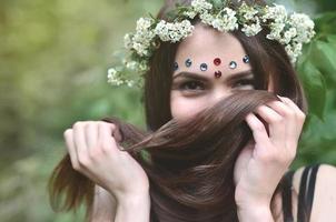 uma foto da floresta de uma bela jovem morena de aparência européia com olhos castanhos escuros e lábios grandes. na cabeça da menina está uma coroa de flores, na testa enfeites brilhantes