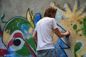 foto no processo de desenho de um padrão de graffiti em uma velha parede de concreto. jovem loiro de cabelos compridos desenha um desenho abstrato de cores diferentes. arte de rua e conceito de vandalismo