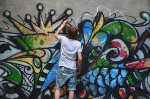 foto de um jovem de short jeans e camisa branca. o cara desenha na parede do grafite um desenho com tintas aerossol de várias cores. o conceito de hooliganismo e danos à propriedade