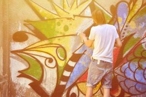 foto de um jovem de short jeans e camisa branca. o cara desenha na parede do grafite um desenho com tintas aerossol de várias cores. o conceito de hooliganismo e danos à propriedade