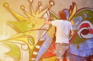 foto de um jovem de short jeans e camisa branca. o cara desenha na parede do grafite um desenho com tintas aerossol de várias cores. o conceito de hooliganismo e danos à propriedade