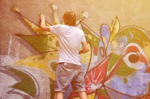 foto de um jovem de short jeans e camisa branca. o cara desenha na parede do grafite um desenho com tintas aerossol de várias cores. o conceito de hooliganismo e danos à propriedade