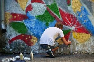 foto no processo de desenho de um padrão de graffiti em uma velha parede de concreto. jovem loiro de cabelos compridos desenha um desenho abstrato de cores diferentes. arte de rua e conceito de vandalismo