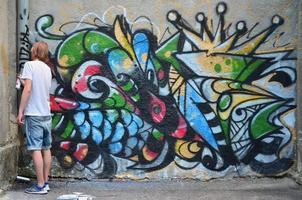 foto no processo de desenho de um padrão de graffiti em uma velha parede de concreto. jovem loiro de cabelos compridos desenha um desenho abstrato de cores diferentes. arte de rua e conceito de vandalismo