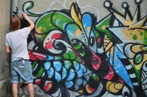 foto no processo de desenho de um padrão de graffiti em uma velha parede de concreto. jovem loiro de cabelos compridos desenha um desenho abstrato de cores diferentes. arte de rua e conceito de vandalismo