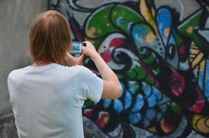 foto no processo de desenho de graffiti em uma velha parede de concreto. um jovem loiro de cabelos compridos tira fotos de seu desenho completo em um smartphone. arte de rua e conceito de vandalismo