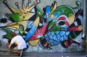 foto de um jovem de short jeans e camisa branca. o cara desenha na parede do grafite um desenho com tintas aerossol de várias cores. o conceito de hooliganismo e danos à propriedade