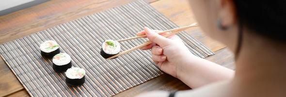 menina morena com pauzinhos detém um rolo de sushi em um fundo de esteira de serwing palha de bambu. comida tradicional asiática foto