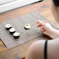 menina morena com pauzinhos detém um rolo de sushi em um fundo de esteira de serwing palha de bambu. comida tradicional asiática foto