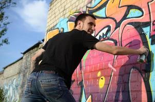 jovem grafiteiro com mochila e máscara de gás no pescoço pinta graffiti colorido em tons de rosa na parede de tijolos. arte de rua e processo de pintura contemporânea foto