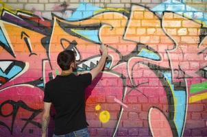 jovem grafiteiro com mochila e máscara de gás no pescoço pinta graffiti colorido em tons de rosa na parede de tijolos. arte de rua e processo de pintura contemporânea foto